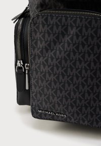 Μαύρη και γκρι τσάντα Michael Kors με επαναλαμβανόμενο μοτίβο λογότυπου "MK", μπροστινή τσέπη, ασημένιο φερμουάρ και μεταλλικό λογότυπο "MICHAEL KORS" στη γωνία στο κάτω μέρος.