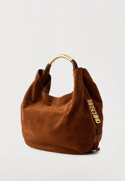 Καφέ τσάντα τύπου hobo από σουέντ με χρυστή μεταλλική λαβή και χρυσό λογότυπο «Moschino» στη μία πλευρά, πάνω σε ανοιχτό φόντο.
