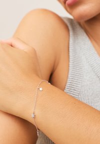 Silver chain bracelet with two star-shaped charms worn on wrist, person wearing a gray sleeveless textured top.