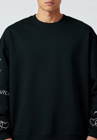 Črna majica s krožnim izrezom, ki ima bele napise in grafike srca na rokavih. Narejena je iz mehke, teksturirane tkanine.