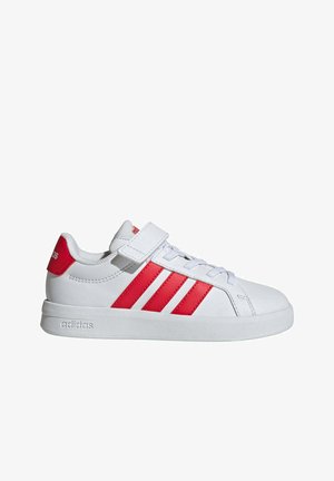 Zapatilla Adidas blanca de corte bajo con rayas rojas, lengüeta roja en el talón, cordones y una tira de velcro sobre fondo blanco.