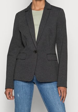 Femme portant un blazer ajusté gris foncé avec un seul bouton et des poches à rabat sur une chemise vert clair et un jean bleu.