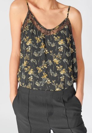 Sort ærmeløs bluse med gul blomsterprint og blonder, båret ind i højtaljede mørkegrå bukser med hænder i lommerne.