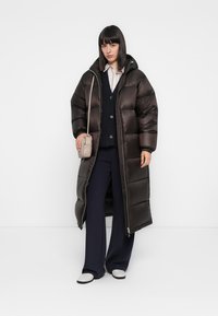 Lange, oversized, bruine pufferjas met een ritssluiting aan de voorkant, capuchon en elastische manchetten. Draagt over een marineblauwe gebreide trui en wijde broek.