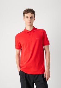 Camisa polo roja de algodón, con cuello, mangas cortas y un logotipo sutil en el pecho. Combinada con pantalones negros.