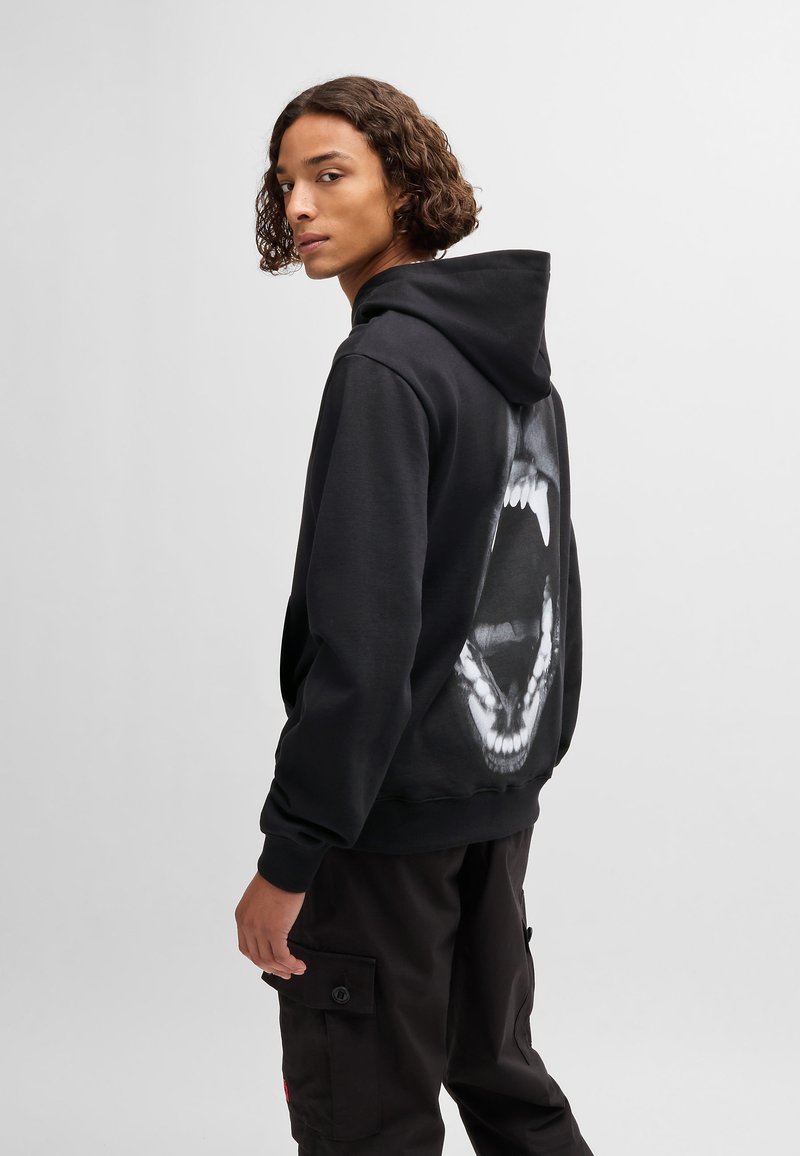 Svart hettegenser med et stort grafisk motiv av en åpen munn på baksiden. Laget av mykt stoff, med snøring på hetten og ribbestrikkede mansjetter.