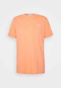 Meruňkově zbarvené tričko s krátkým rukávem od Adidas s kulatým výstřihem a malým bílým logem Adidas na levém hrudníku.