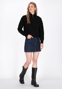 Zwarte gebreide trui met halve rits, gecombineerd met een denim mini-jurk. Zwarte kniehoge laarzen met gespaccenten maken de outfit compleet.