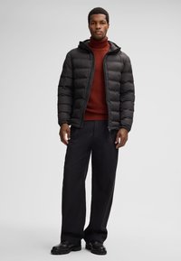 Giacca imbottita nera con zip frontale, cappuccio e trama trapuntata, indossata sopra un maglione rosso lavorato a maglia, abbinata a pantaloni neri a gamba larga e stivali.