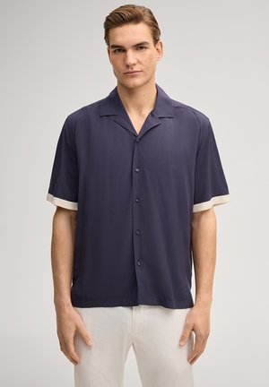 Junger Mann trägt ein kurzärmeliges marineblaues Hemd mit weißen Manschetten und weiße Hosen, steht vor einem hellgrauen Hintergrund.