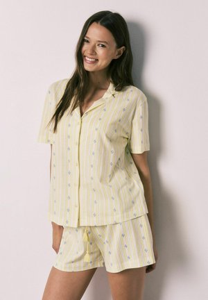 Femme souriante portant une chemise à manches courtes jaune clair avec boutons et un short assorti avec des rayures verticales et de petits motifs floraux bleus.