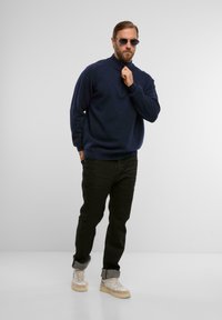 Marineblauer Pullover mit hohem Kragen, kombiniert mit schwarzen Slim-Fit-Jeans und beigen Sportschuhen mit strukturiertem Design und weißen Akzenten.