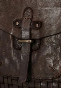 Détail d'un sac en cuir marron avec une sangle centrale et une boucle en métal, des accents cousus et un fond tissé texturé.