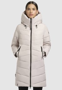 Manteau rembourré beige clair avec un col montant, une capuche à cordon de serrage et une fermeture éclair. Présente des coutures diagonales et deux poches latérales.