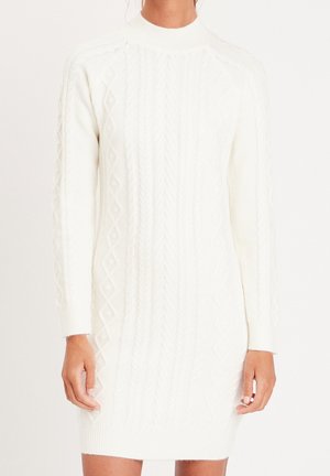Femme portant une robe-pull blanche à manches longues en maille torsadée avec un col montant, montrée du cou jusqu'à mi-cuisse sur un fond uni.