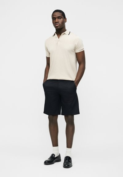 Homme debout, les mains dans les poches, portant un polo beige à manches courtes, un short noir, des chaussettes blanches et des mocassins noirs sur un fond uni.