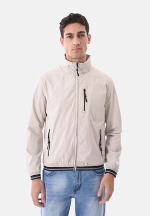 Uomo che indossa una giacca beige chiaro con zip, tasche nere con cerniera e polsini a righe, abbinata a jeans blu, in piedi davanti a uno sfondo bianco.