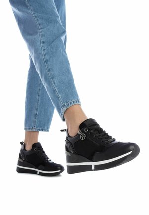 Zapatillas cuña negras con tela texturizada y detalles en cuero, con un zipper lateral y herrajes metálicos decorativos, combinadas con denim claro.