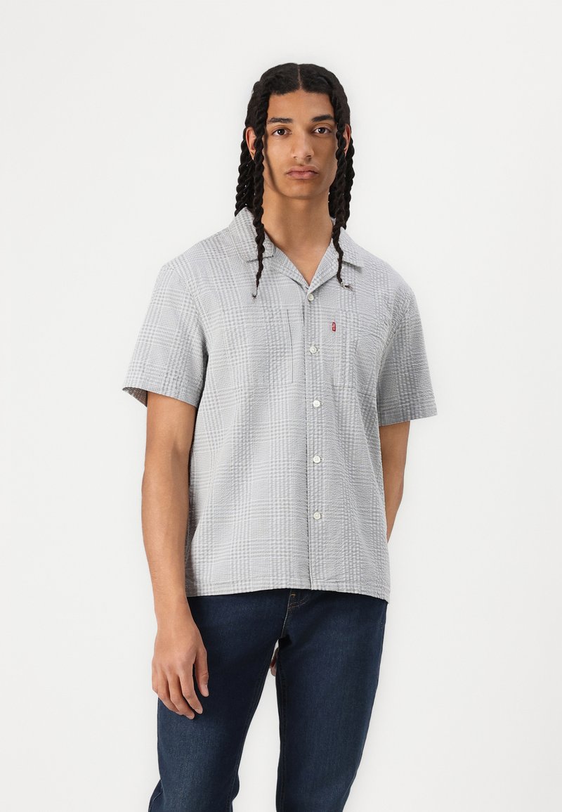 Chemise à manches courtes en tissu léger et texturé de couleur grise avec un motif à carreaux subtil. Dotée d'un col pointu et d'une fermeture à boutons.