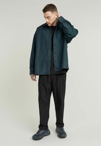 Tealfärgad skjorta med knappar och en enda bröstficka, bärs över en svart skjorta, i kombination med lösa svarta byxor och kraftiga grå sneakers.