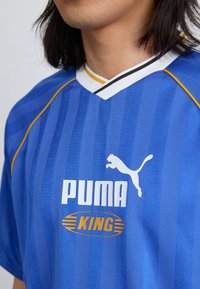 Modrá športová dres s vertikálnymi pruhmi, bielym golierom a žltými a čiernymi akcentmi. Obsahuje logo 'PUMA' a text 'KING' v zlatej farbe.