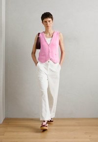 Gilet rosa lavorato a maglia con bottoni, pantaloni bianchi a righe e sneakers bordeaux. Una borsa a tracolla aggiunge un tocco al look casual dell'outfit.
