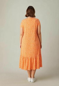 Ein oranges Kleid mit floralem Muster, kurzen Capärmeln und einem gestuften Rüschensaum. Aus leichter Stoffqualität, knielang. Weiße Schuhe sind sichtbar.
