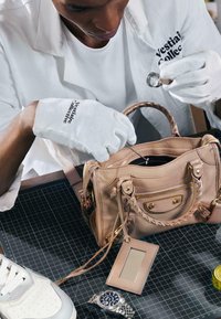 Personne portant des gants blancs examinant un sac à main beige avec une loupe et un outil sur une surface noire quadrillée.