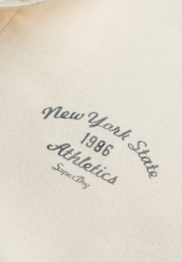 Hvidt stof med grå tekst, der lyder "New York State 1986 Athletics Super Dry" i buet og skråskrift.