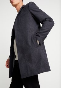 Šedý vlněný kabát s klopami s výřezem, bočními kapsami a tříknopovým zapínáním vpředu, s hladkou texturou a střihovou úpravou.