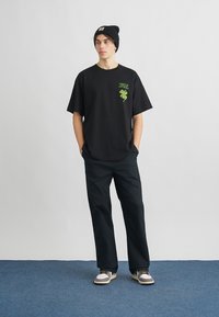 Czarna bawełniana koszulka z zielonym nadrukiem koniczyny na piersi, zestawiona z ciemnymi spodniami i czarną czapką typu beanie. Luźny krój, minimalne detale.