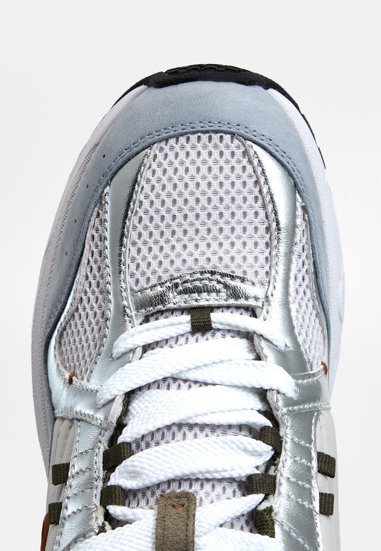 Sneaker met een bovenwerk van gaas in wit en zilver, met textuuraccenten, reflecterende details en witte veters. De zool is zwart.