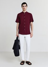Chemise bordeaux à manches courtes avec patte de boutonnage, associée à un pantalon slim blanc et des sandales marron à enfiler, tenant un sac cabas noir.