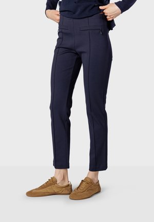 Personne portant un pantalon ajusté bleu foncé avec des poches zippées à l'avant et des baskets marron décontractées, debout devant un fond uni.