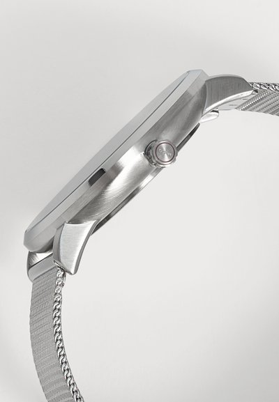 Vue latérale d’une montre-bracelet argentée avec un bracelet en maille métallique et une couronne ronde sur le côté droit du boîtier de la montre.