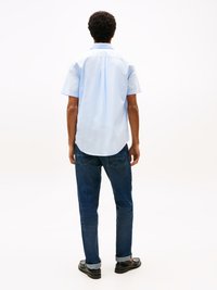 Homem com cabelo encaracolado, vestindo uma camisa de manga curta azul clara, jeans azul escuro e mocassins pretos, de pé, de costas, em um fundo branco.