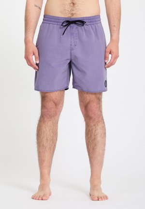 Hombre con pantalones cortos de baño morados hasta la mitad del muslo con cordón negro, de pie descalzo contra un fondo blanco.