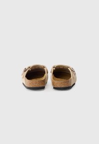 Kreminės spalvos slip-on tipo klumpės su minkštu viršumi, kamštiniu padu ir tekstūruotu juodu padu. Papuoštos dekoratyviniu metaliniu sagtimi.