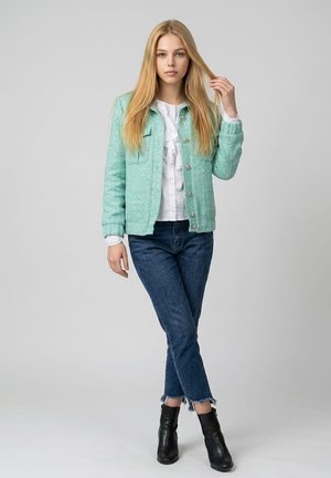Joven mujer con cabello rubio largo que lleva una chaqueta verde menta, blusa blanca, vaqueros azules y botas negras hasta el tobillo, de pie frente a un fondo gris.