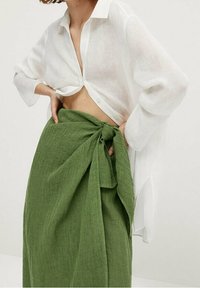 Chemise blanche en lin à boutons avec des manches retroussées, associée à une jupe en lin vert avec un détail de taille nouée et un tissu texturé.