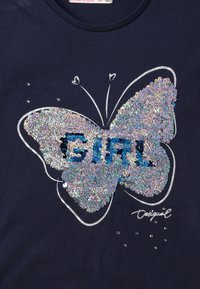 Námořnické tričko s velkým, blyštivým designem motýla vyrobeným z flitrů. Text "DÍVKA" je modrý, obklopený malými kamínky a obrysy.