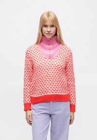 Pletený sveter s ružovým vysokým golierom a telom s oranžovým, bielym a červeným vzorom. Rebrované manžety a lem, mäkká textúra. Kombinovaný s svetlofialovými nohavicami.
