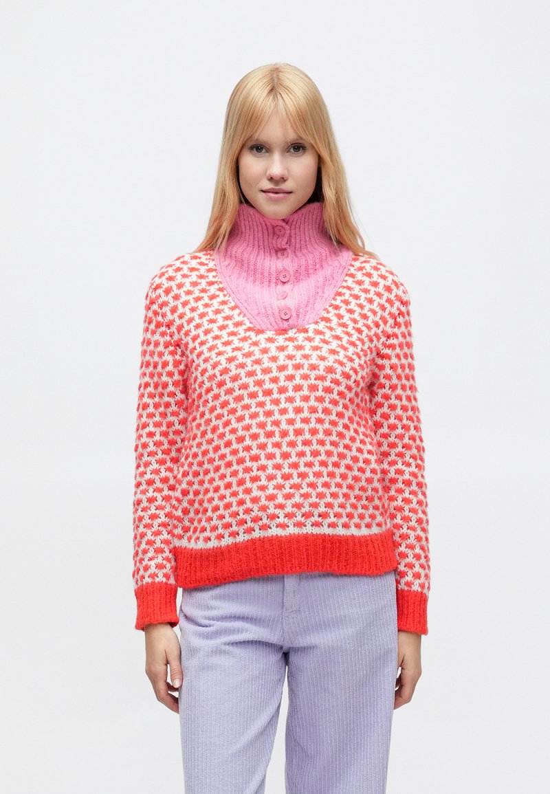 Pletený sveter s ružovým vysokým golierom a telom s oranžovým, bielym a červeným vzorom. Rebrované manžety a lem, mäkká textúra. Kombinovaný s svetlofialovými nohavicami.