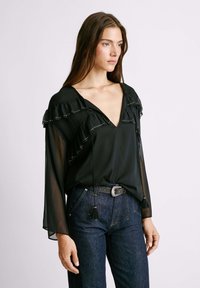 Blusa negra con mangas transparentes y acampanadas, volantes en el escote y acentos de tachuelas metálicas. Combinada con unos jeans de textura y un cinturón decorativo.