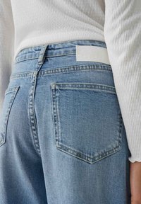 Primo piano di una persona che indossa jeans in denim blu chiaro e una maglia bianca a maniche lunghe con trama, con particolare attenzione alla tasca posteriore e all'area della vita.
