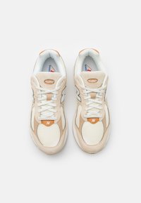 Béžové sneakers New Balance s vrchní částí z síťoviny a semiše, bílé tkaničky a oranžový akcent na jazyku; obsahují logo a polstrovaný límec.