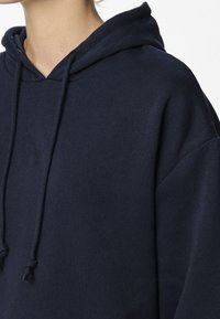 Marinblå hoodie med dragsko, tillverkad av mjukt tyg. Har en stor huva och en lös passform med sänkta axlar för komfort.