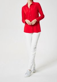 Chemise rouge à manches longues avec un design à boutons, associée à un pantalon blanc ajusté et des baskets claires. Texture de tissu lisse, look décontracté.