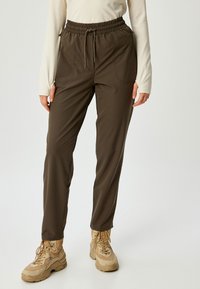 Braune Stretchhose mit elastischem Bund und zwei Vorderaschen, gerades Bein-Design und einem leichten Stoff. Kombiniert mit hellbraunen Wanderschuhen.