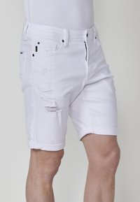Pantalones cortos de mezclilla blanca con cremallera, que presentan detalles desgastados en la parte delantera, dobladillos remangados y un diseño clásico de cinco bolsillos.
